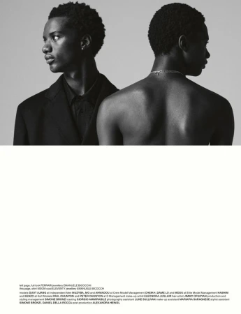 Two dark-skinned male models pose back to back, one wearing a black suit, the other shirtless with a silver chain. Black and white portrait.