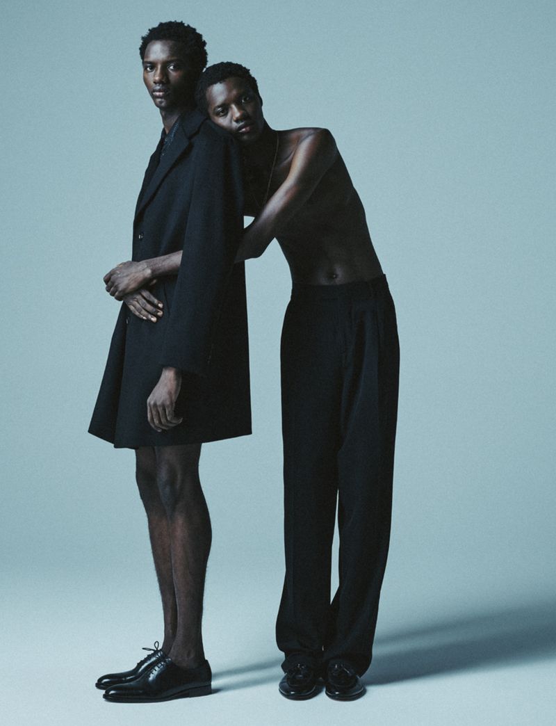Two black male fashion models pose back-to-back in a studio with a pale blue backdrop, one wearing a black coat and shoes, the other in pants and a top.