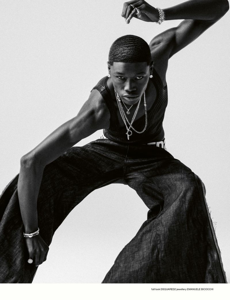Fashion shot of a Black male model in a sleeveless top and wide-leg jeans, accessorized with layered necklaces and bracelets, posing dynamically.