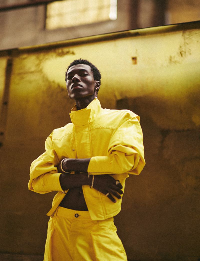 A Black male model poses in a yellow jacket and matching pants in an industrial indoor setting, his arms crossed, conveying confidence.
