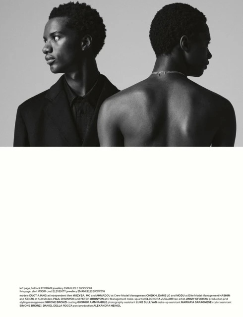Black-and-white portrait of two men, one in a black blazer and shirt, and another shirtless from behind with a silver necklace.