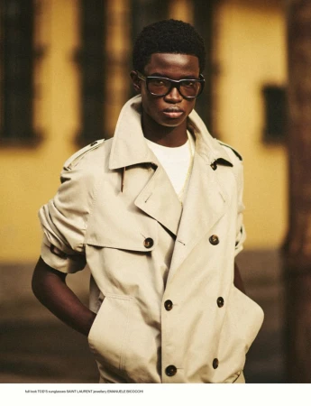 Black male model wearing a beige double-breasted trench coat and dark sunglasses against an out-of-focus yellow building exterior