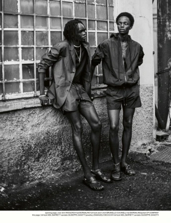 Monochrome editorial fashion photograph of two male models standing together outside against an industrial wall