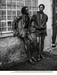 Monochrome editorial fashion photograph of two male models standing together outside against an industrial wall