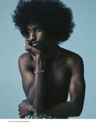 Portrait of a shirtless Black man with a large afro resting his chin on his hand against a plain grey-blue studio backdrop.