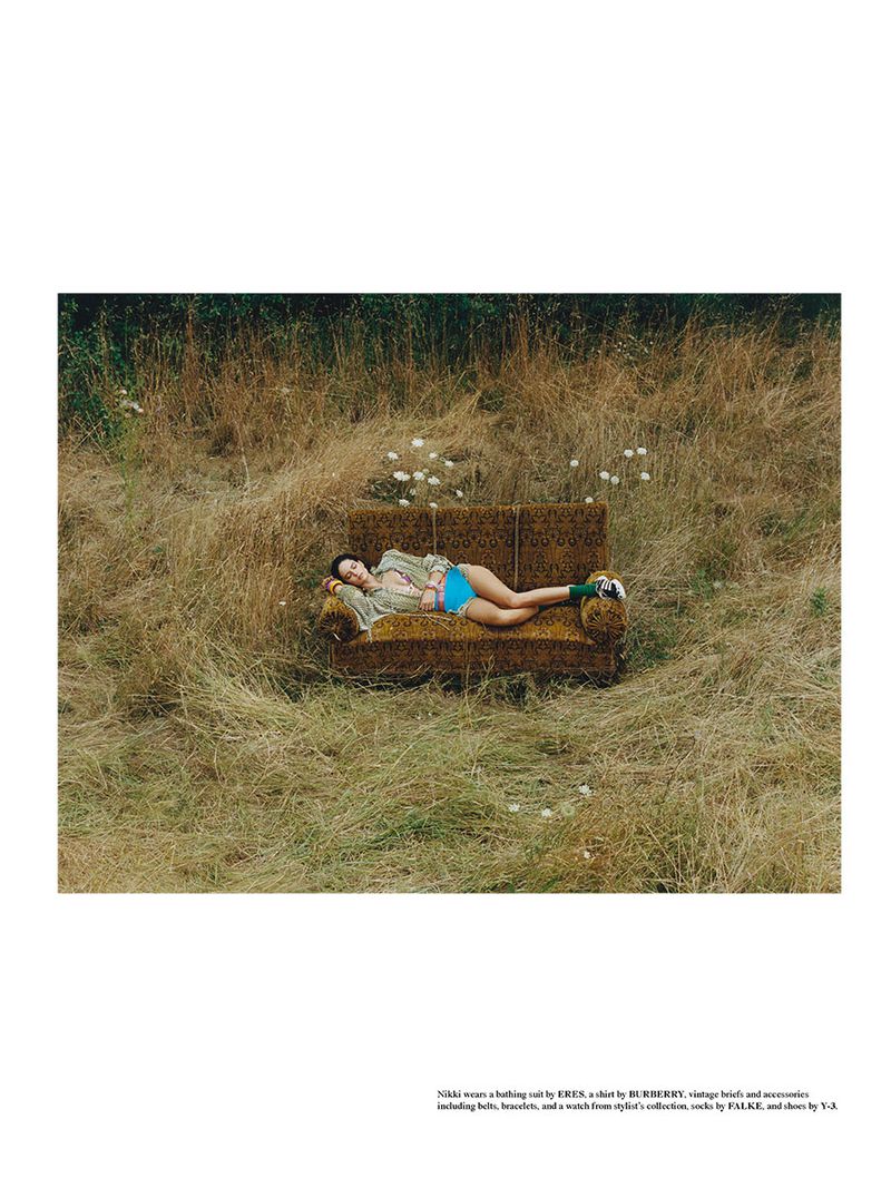 Woman in bathing suit and shirt lies on couch outdoors in dry grassy field with wildflowers in a fashion editorial shoot.