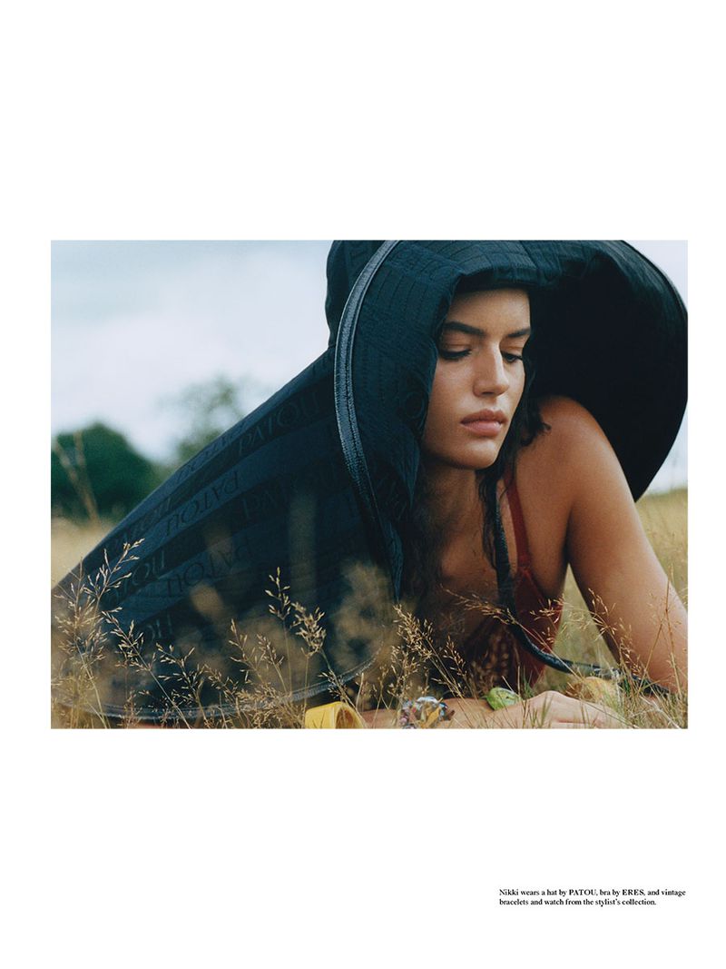 Portrait of a woman in a black hat lying in a field of tall grass, wearing a reddish-brown top, outdoors on a cloudy day.
