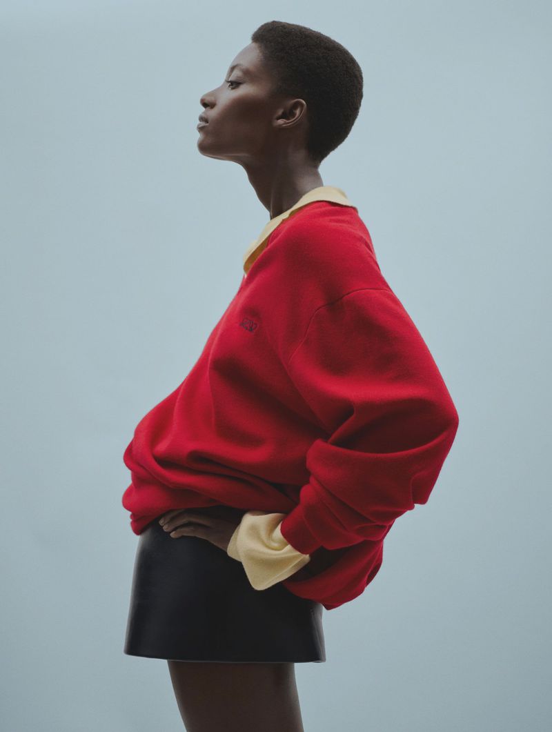 Profile shot of an African American adult woman in a red sweatshirt and black skirt against a light blue backdrop in a fashion editorial.