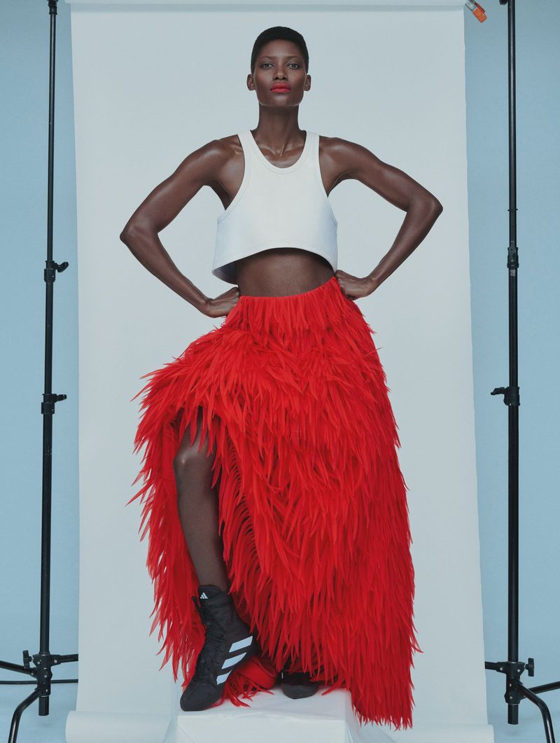Fashion shot of a Black woman posing in a white crop top, and a long red fringe skirt. She wears sneakers in a light studio.