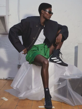 Dark-skinned woman wearing black jacket, green shorts, sunglasses, and Adidas boxing shoes, posing in an indoor setting with wood floor.
