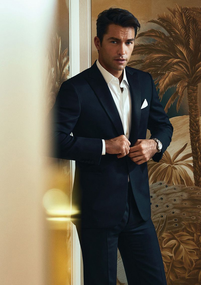 Man in a black suit with a white pocket square buttons up his suit jacket in a neutral-toned room, with a palm tree design behind him.