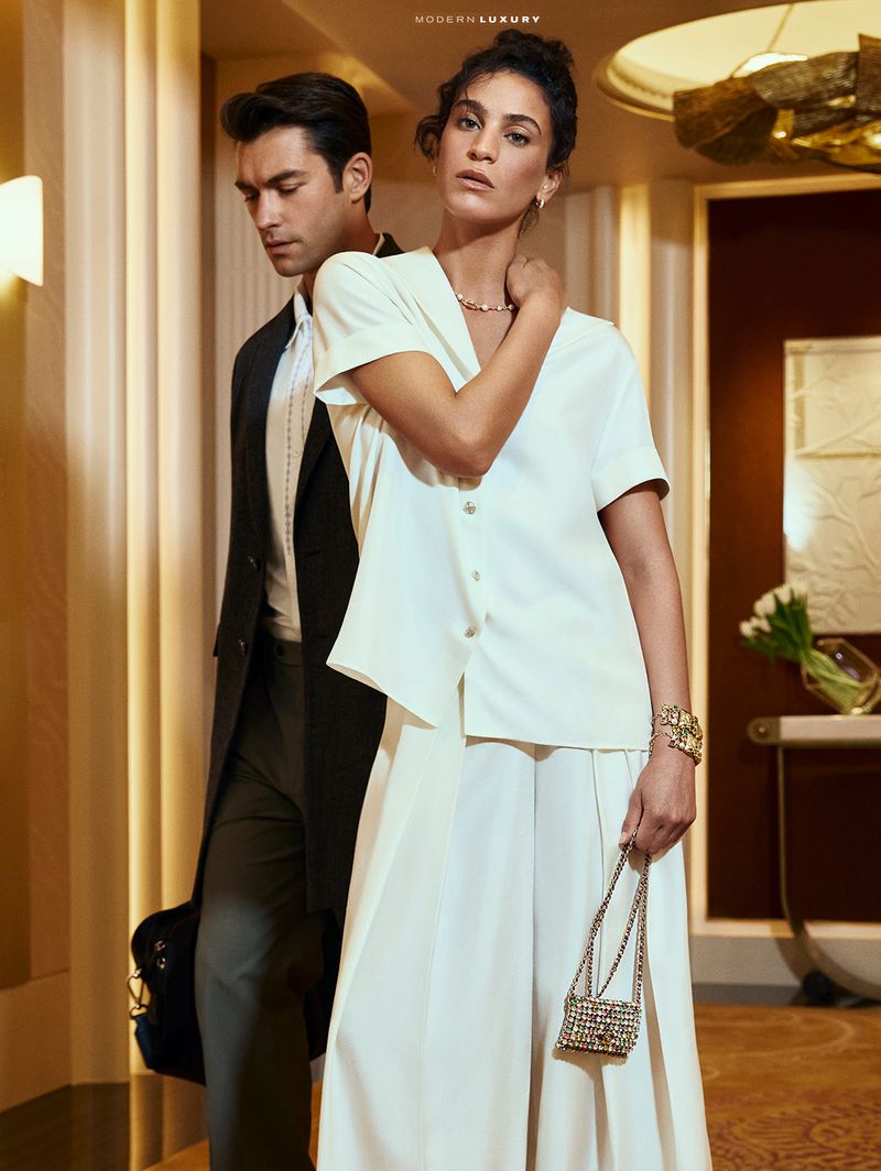Elegant couple in a modern hotel hallway. The woman wears a white blouse and culottes, carrying a beaded bag. The man wears a dark suit.