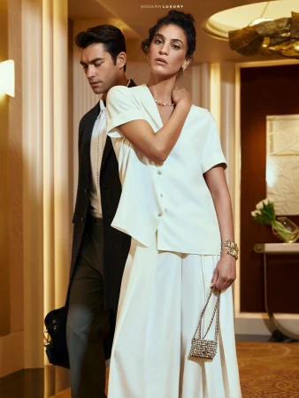 Elegant couple in a modern hotel hallway. The woman wears a white blouse and culottes, carrying a beaded bag. The man wears a dark suit.