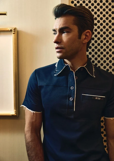 A handsome adult male poses indoors wearing a dark blue collared shirt with white trim, styled hair, and neutral expression looking to the side.