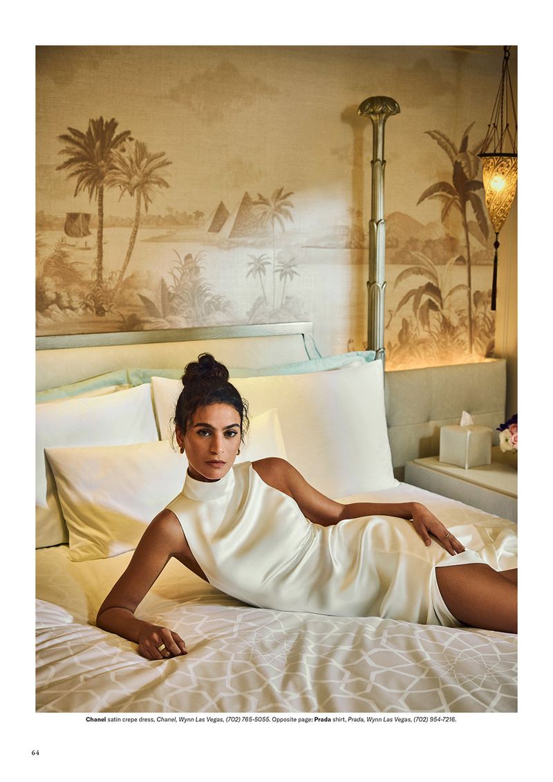Adult woman in a dress poses on a patterned bed with pillows and a headboard in front of a tan mural in a light-filled interior.