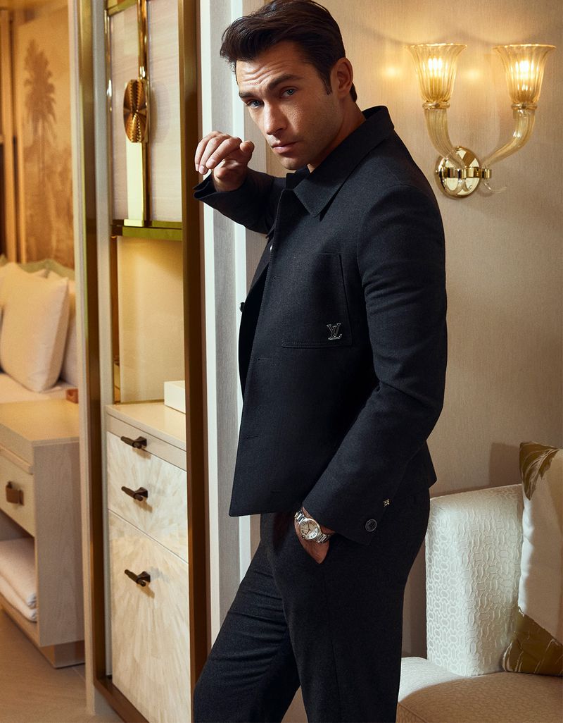 Man in a black suit and wristwatch poses indoors next to a cabinet and decorative wall lights, exuding a sophisticated, stylish vibe.