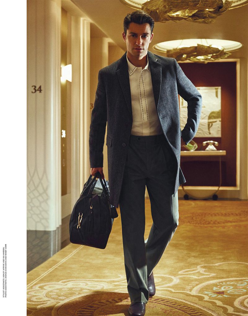 Stylish man in a suit walks through a hotel hallway with a duffel bag, showcasing classic fashion in an elegant interior setting.