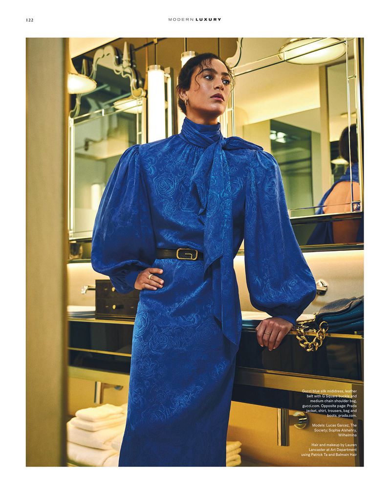 Fashion editorial shot of a woman wearing a blue high-necked, belted dress with puffed sleeves in a bathroom with gold fixtures.