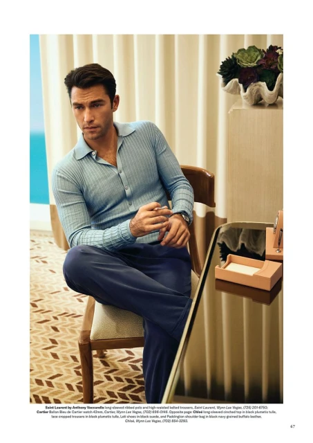 An adult male poses indoors, wearing a light blue collared shirt and navy pants, sitting on a chair with a table nearby.