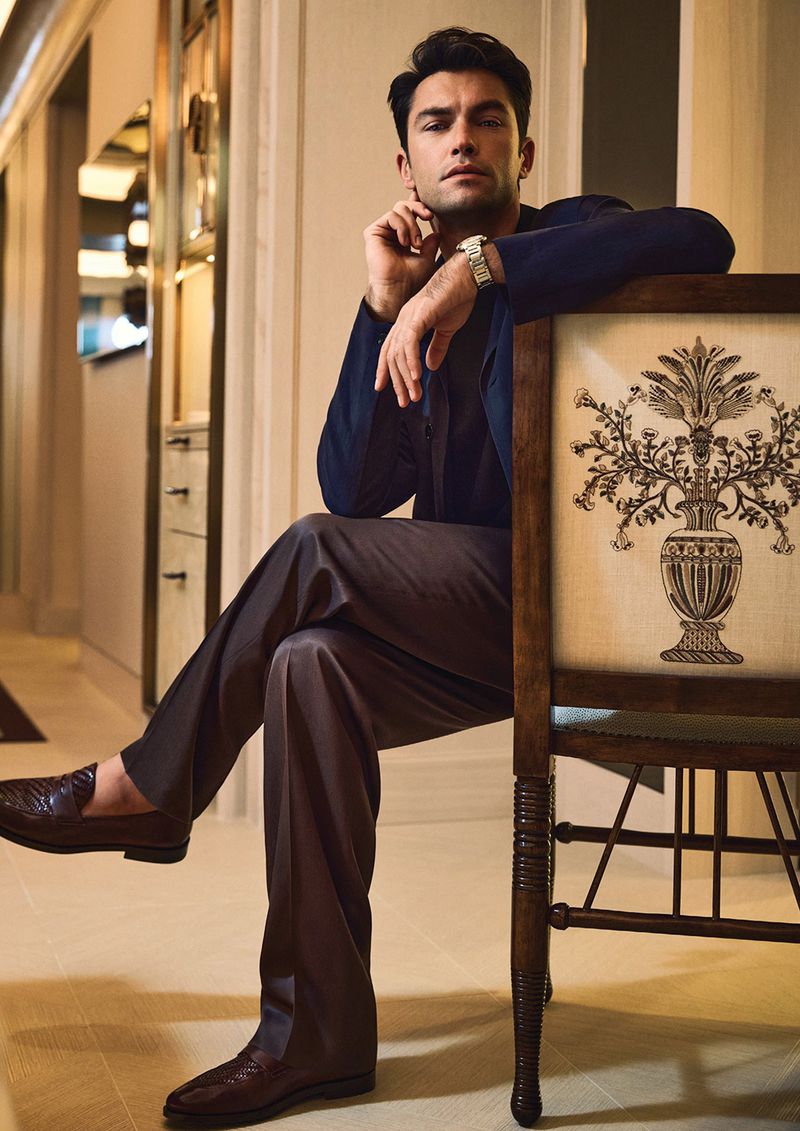 Adult man seated in elegant room with dark suit, brown trousers, and dress loafers; decorative chair and stylish interior visible.