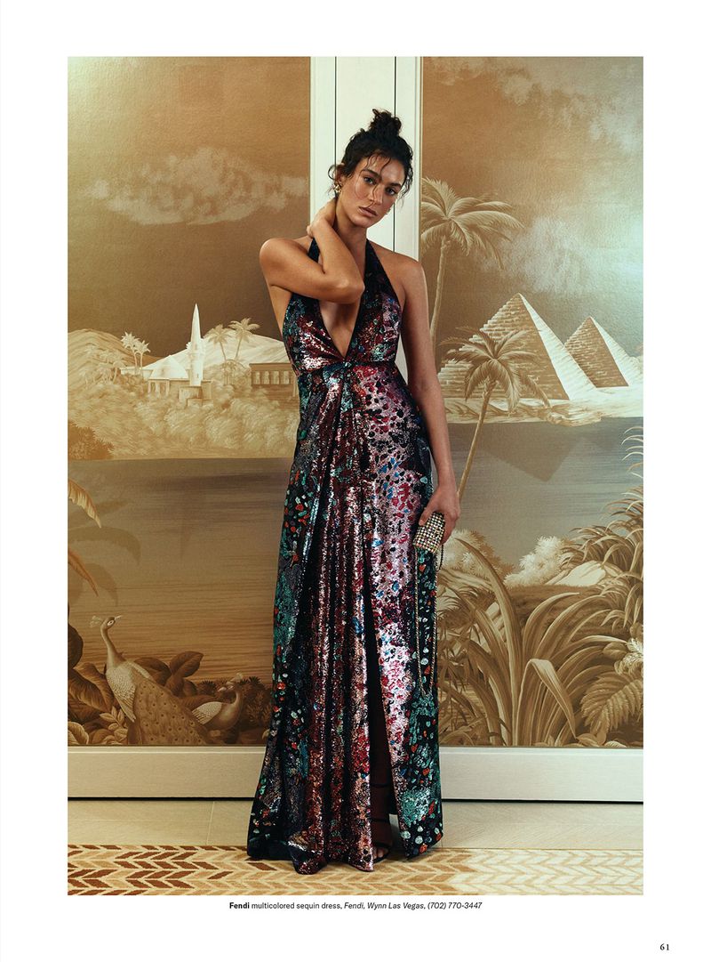 Woman in a multicolored halter gown stands in front of a scenic wallpaper featuring pyramids and palm trees in an editorial fashion shot.