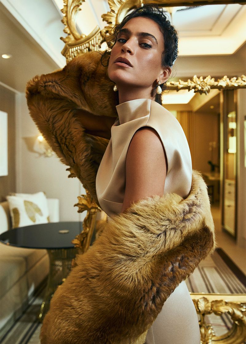 Elegant woman in a beige dress poses indoors with a fur stole and elaborate gold-trimmed mirror in a beauty or fashion editorial.