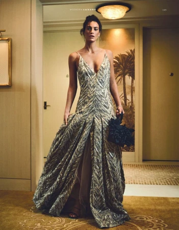 Woman in elegant gown poses indoors, holding a black clutch, with a framed palm tree artwork in the background, bathed in warm lighting.