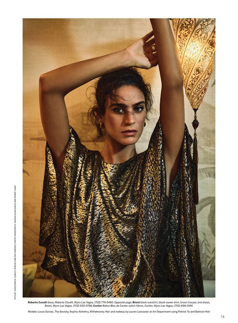 Woman poses in front of a patterned wall with an ornate lamp. She wears a gold, patterned kaftan, her arms raised overhead.