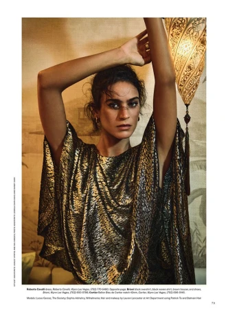Woman poses in front of a patterned wall with an ornate lamp. She wears a gold, patterned kaftan, her arms raised overhead.