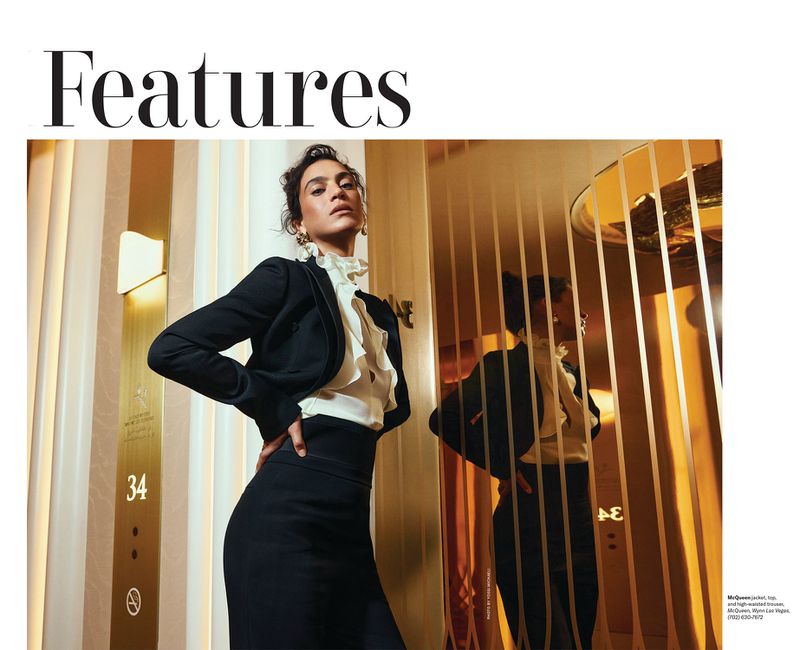 Elegant woman in a black skirt suit poses in front of a vertically striped gold mirrored wall in a fashion editorial style photograph.