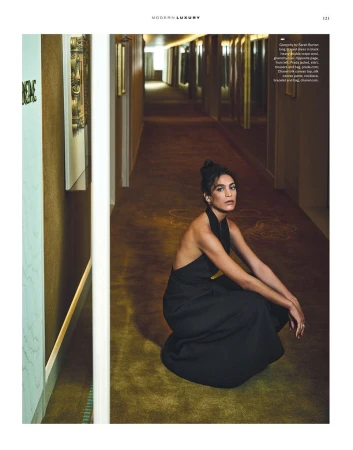 A woman in a black dress poses while crouching in a hotel hallway with carpet and paneled walls, looking at the camera.