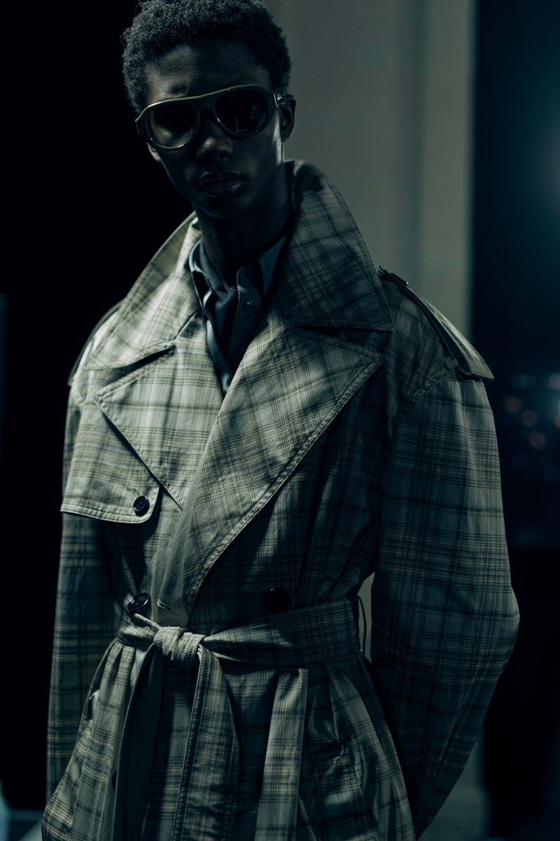 A stylish person wearing a plaid trench coat and aviator sunglasses stands in front of a dark background, striking a fashionable pose.