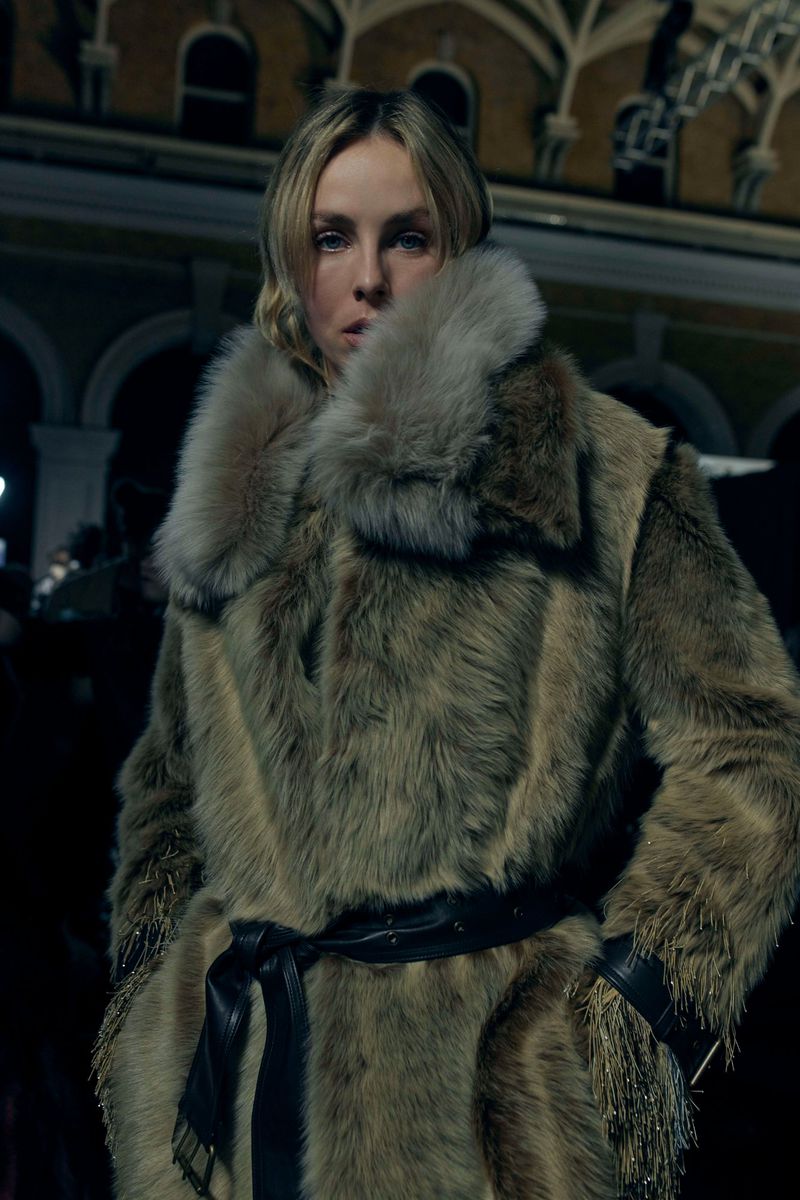Blonde woman in a furry coat with a black belt stands in front of an arched building with outdoor lighting at dusk.