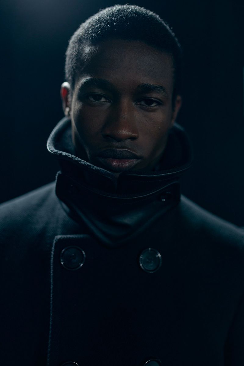 Portrait of an African American man in a double-breasted overcoat with a wide collar against a dark background. Moody lighting and close framing.
