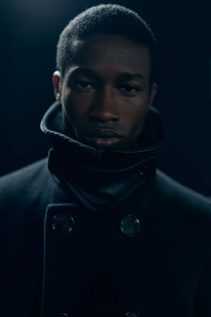 Portrait of an African American man in a double-breasted overcoat with a wide collar against a dark background. Moody lighting and close framing.