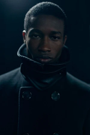 Portrait of an African American man in a double-breasted overcoat with a wide collar against a dark background. Moody lighting and close framing.