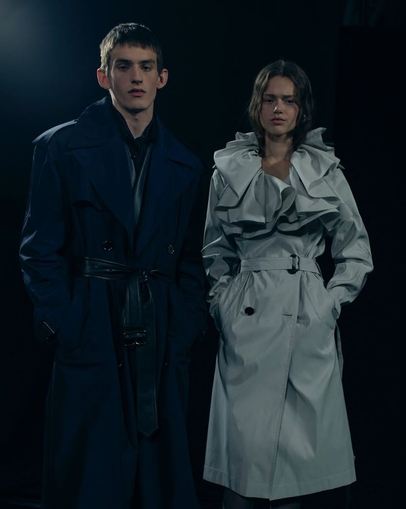 Two models stand in a dark studio showcasing contemporary trench coats, one in dark blue, the other in light gray with ruffle detail.