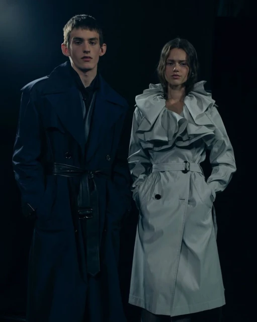 Two models stand in a dark studio showcasing contemporary trench coats, one in dark blue, the other in light gray with ruffle detail.