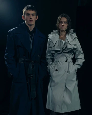 Two models stand in a dark studio showcasing contemporary trench coats, one in dark blue, the other in light gray with ruffle detail.