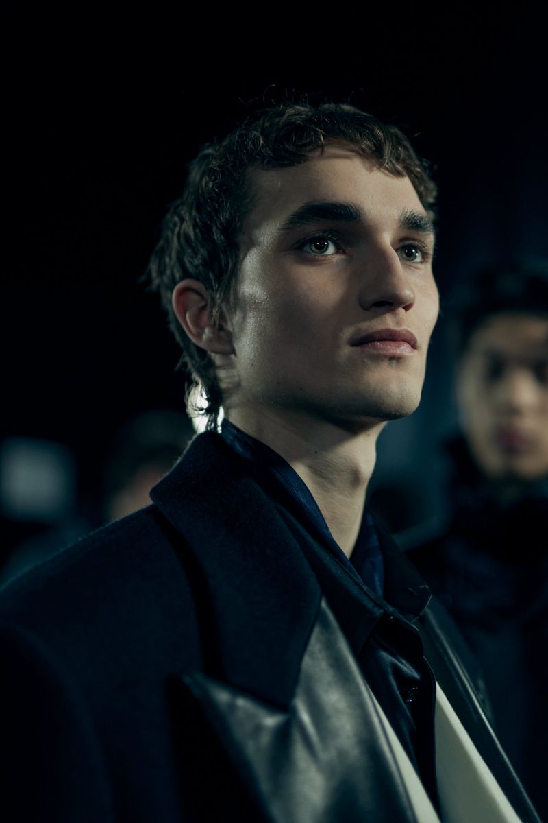 Close-up of a young male model with dark curly hair in a black coat looking up against a dark backdrop with another person in soft focus.