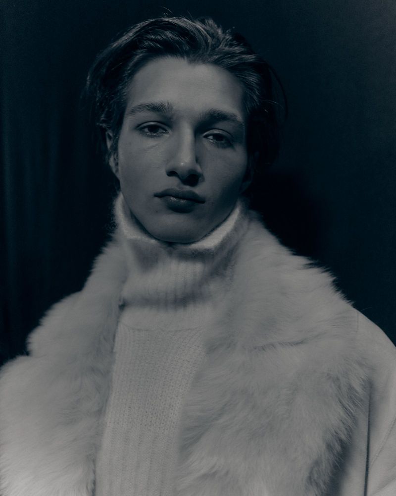 Portrait of a young man wearing a white turtleneck sweater and a faux fur coat against a dark background in a studio setting.