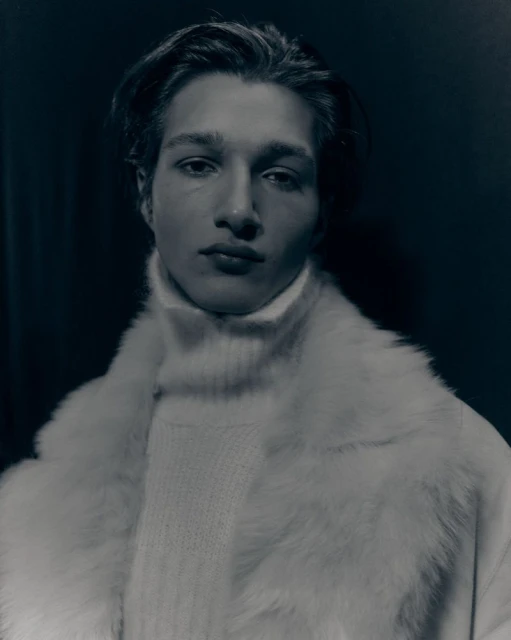 Portrait of a young man wearing a white turtleneck sweater and a faux fur coat against a dark background in a studio setting.