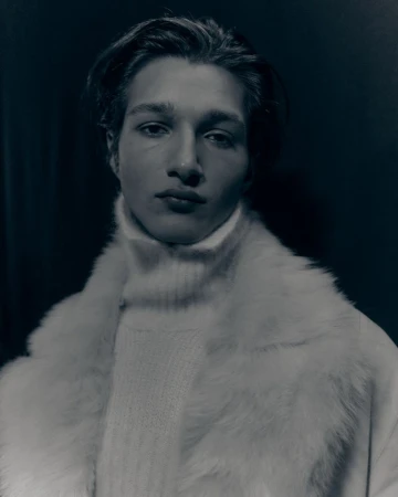 Portrait of a young man wearing a white turtleneck sweater and a faux fur coat against a dark background in a studio setting.