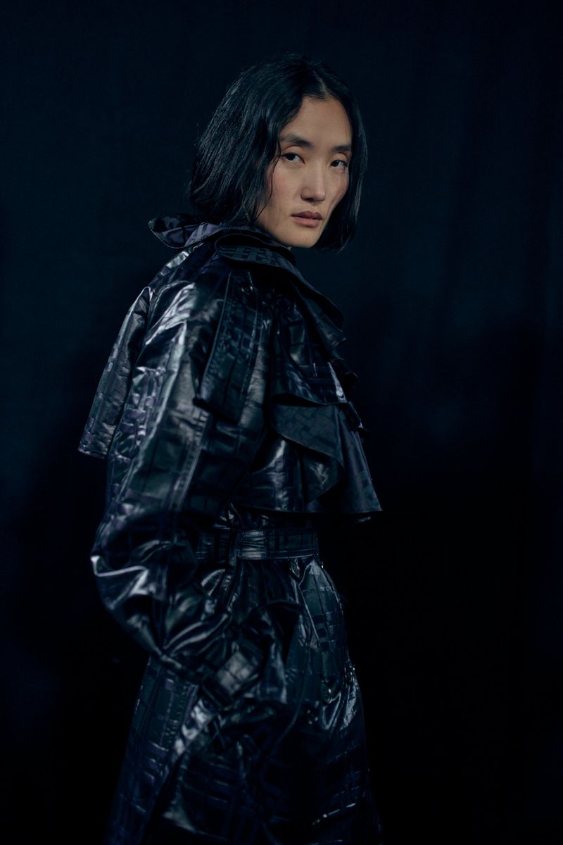 Portrait of an adult woman with short black hair wearing a shiny, black vinyl trench coat in a studio setting with dark backdrop.
