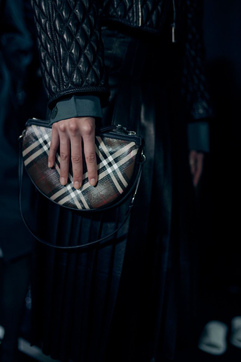 A woman holds a Burberry handbag with a plaid pattern, showcasing fashion details like a quilted leather jacket in a dimly lit setting.