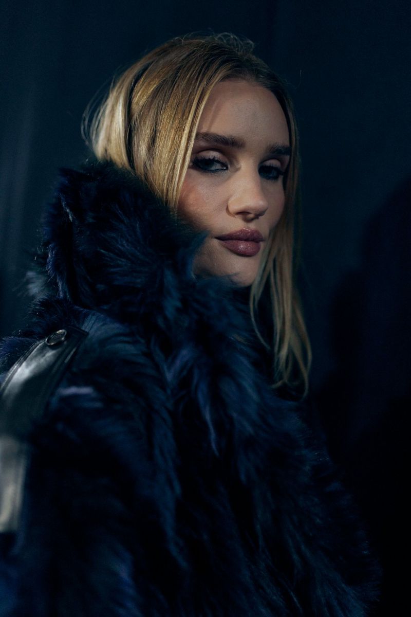 Close-up portrait of a blonde adult woman with dark eye makeup, wearing a blue fur coat in an indoor setting with dark lighting.