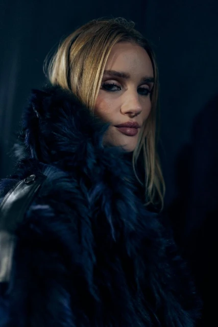 Close-up portrait of a blonde adult woman with dark eye makeup, wearing a blue fur coat in an indoor setting with dark lighting.