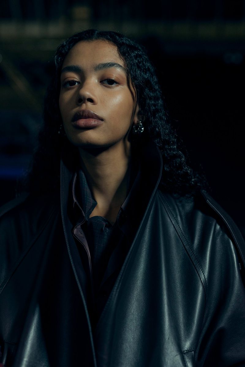 Portrait of a woman with dark hair and a black leather coat, with an eye-level angle and shallow depth of field in a dimly lit indoor setting.