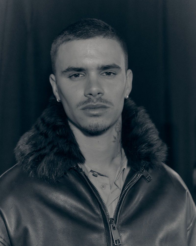 Black and white portrait of a young adult man in a leather jacket with fur collar, looking directly at the camera in a dark indoor space.