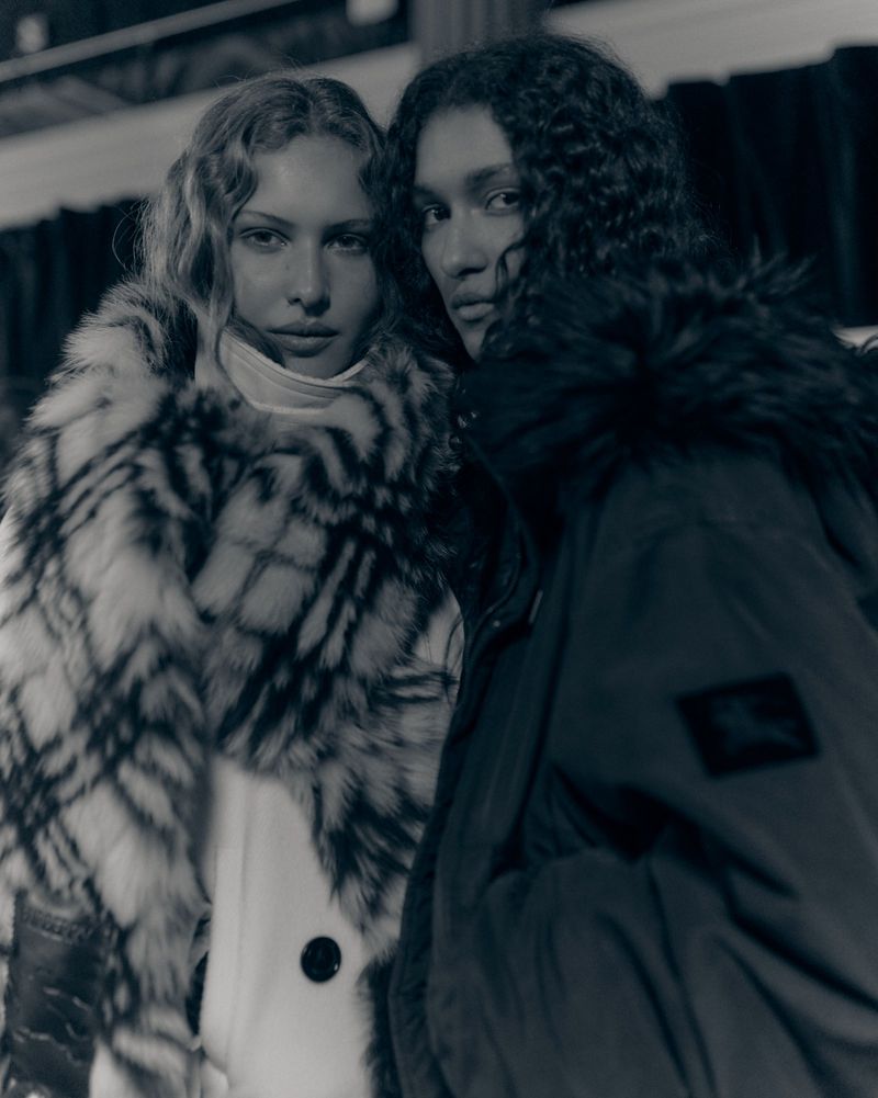 Two adult women pose indoors, one in a patterned faux fur coat, the other in a dark parka, both looking at the camera in monochrome.